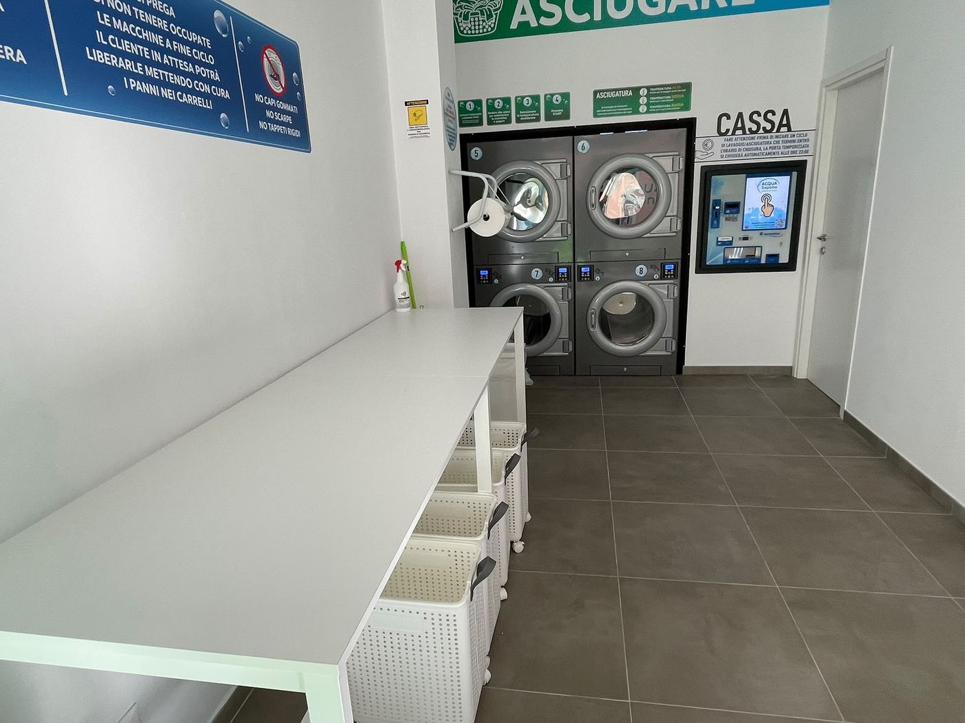 Interno luminoso della lavanderia self-service Acqua Sapone a Pontecurone, con asciugatrici, cassa automatica e tavolo per piegare il bucato